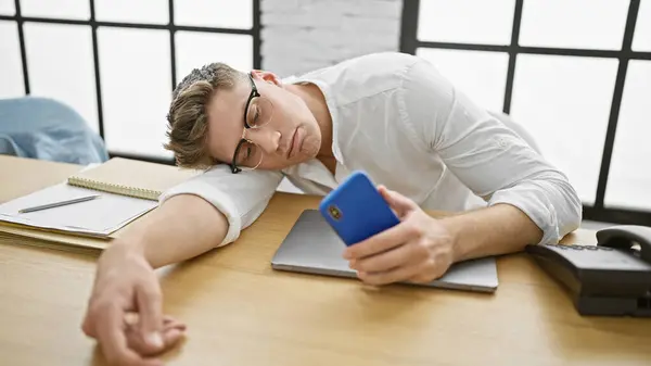 精疲力竭的年轻白种人商人 手里拿着智能手机 与睡在办公室桌子上的压力作斗争 战战战兢兢 工作单调乏味 — 图库照片