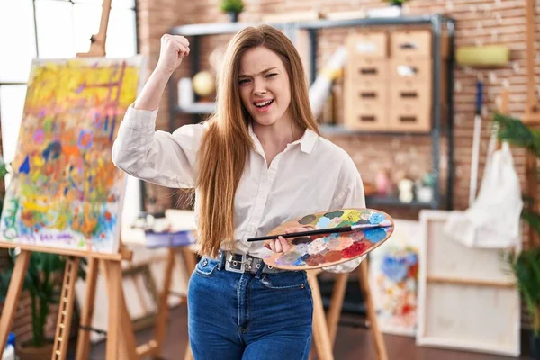 Young caucasian woman at art studio holding palette annoyed and frustrated shouting with anger, yelling crazy with anger and hand raised