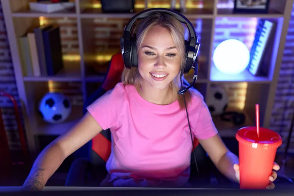 Young blonde woman streamer playing video game drinking beverage at gaming room
