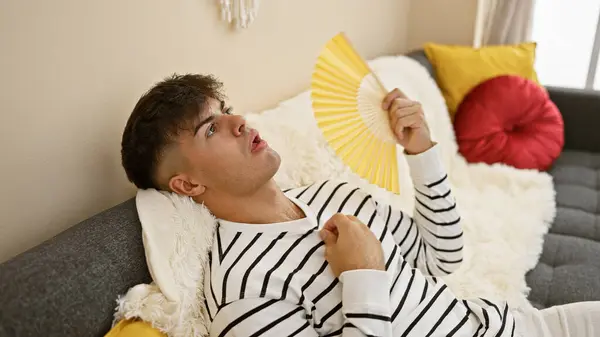 Handsome young hispanic man sweating from the heat, finds comfort in the cool breeze of a hand fan, sitting on a cozy sofa in the living room at home, worry etched on his face.