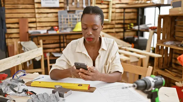 Serious african american female carpenter proficiently texting on smartphone while gracefully sitting at carpentry workshop table