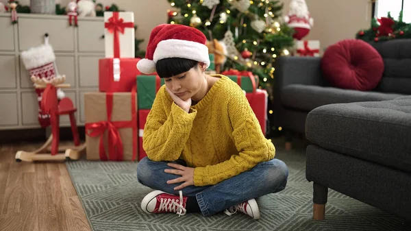 Young chinese woman sitting by christmas tree with boring expression at home