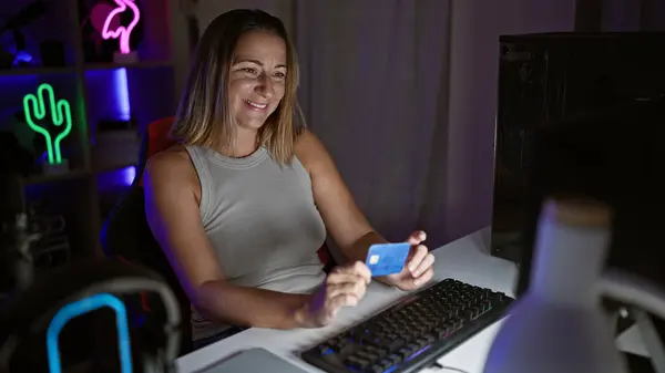 Vibrant, young blonde streamer girl live-streaming gaming session from cozy home office, smiling as she uses credit card on her computer in the heat of the night