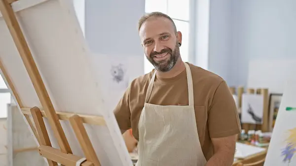 Attractive young man, confident artist, smiling while drawing in art studio, enjoys hobby in apron with paintbrushes