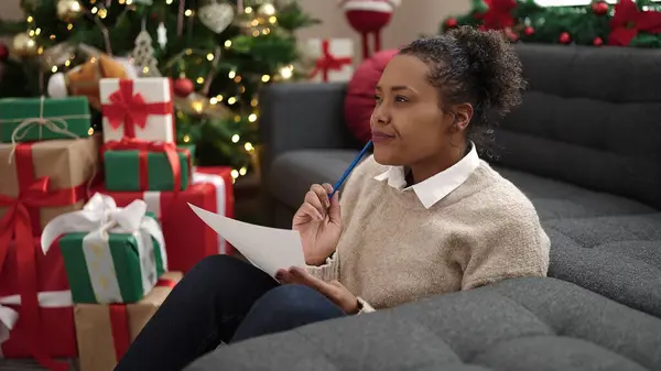 African american woman writing on santa claus letter sitting on floor by christmas tree at home