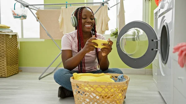 Joyful african american woman multitasking in the laundry room, playing a video game while smiling and washing clothes