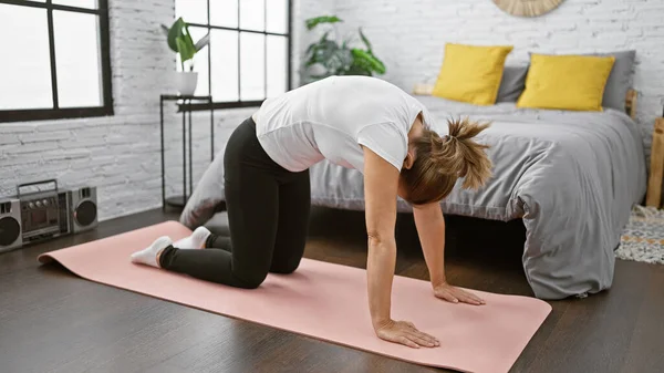 Çekici, spor giyimli genç sarışın kadın sabah dengesini yatak odasında dinlendirici bir sırt esnemesiyle buluyor. Kırışık yatağının yanında, gülümsüyor ve gündüze doğru esniyor..