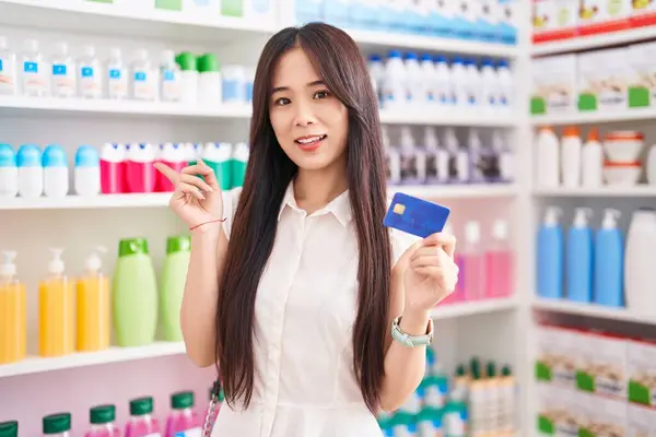 年轻的中国女人在药店购物 手里拿着信用卡 面带微笑 手指手画脚地朝旁边走去 — 图库照片