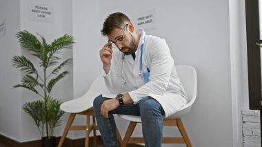 Aşırı çalışan İspanyol asıllı genç bir adam, üniformalı stresli bir doktor kliniğin bekleme odasında ciddi bir şekilde oturuyor, sağlık hizmetleri stresinin bedelini gösteriyor.