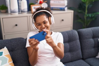 Evdeki kanepede oturan Afrikalı Amerikalı kadın video oyunu oynuyor.