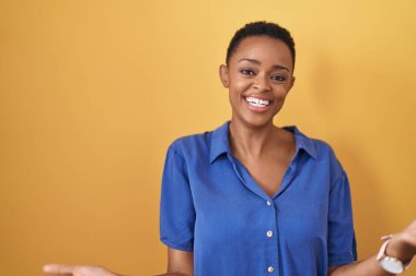 Sarı arka planda duran Afro-Amerikalı kadın gülümseyerek gülümsüyor. Dostça bir hoş geldin, olumlu ve kendinden emin selamlar. 