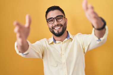 İspanyol genç adam iş kıyafetleri ve gözlük takmış kameraya bakıyor kucaklaşmak için kollarını açmış gülümsüyor. Mutluluğu kucaklayan neşeli bir ifade.. 