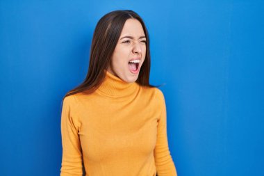 Genç esmer kadın mavi arka planda kızgın ve kızgın bağırıyor sinirli ve öfkeli, öfkeyle bağırıyor. öfke ve agresif konsept. 