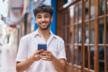 Genç Arap adam sokakta akıllı telefon kullanarak kendinden emin gülümsüyor.