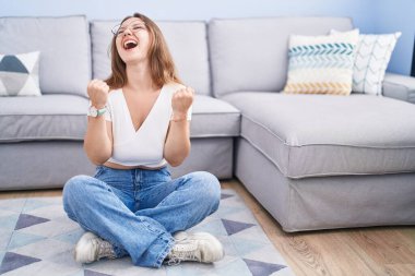 Oturma odasında yerde oturan beyaz bir kadın kolları havada ve gözleri kapalı bir şekilde başarıyı kutluyor ve şaşırıyor. Kazanan konsept. 