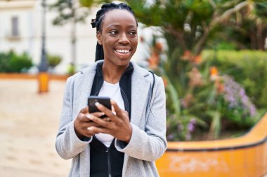 Afro-Amerikalı kadın parkta akıllı telefon kullanarak gülümsüyor.