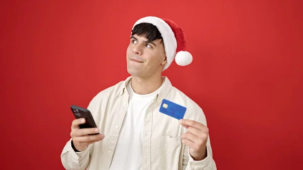 スマートフォンとクレジットカードで買い物をする若いヒスパニック系男性は 隔離された赤い背景の上にクリスマスの帽子をかぶっています — ストック写真