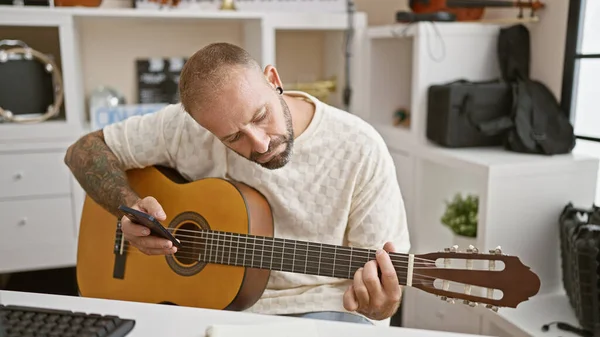 Attractive young man, relaxing in the music studio, playing classic tunes on guitar, composing via smartphone