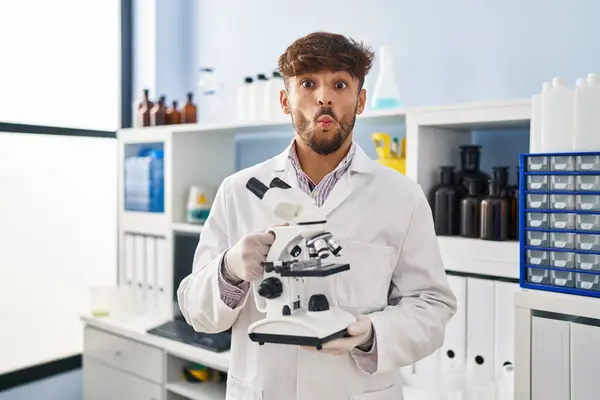 Arab man with beard working at scientist laboratory holding microscope making fish face with mouth and squinting eyes, crazy and comical.