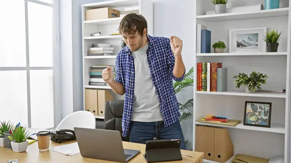 Joyful young business man celebrating work success at office desk, confidently working online with his laptop
