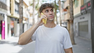 Yakışıklı, yakışıklı İspanyol bir adam akıllı telefonuyla ciddi ciddi sohbet ediyor. Güneşli şehir sokağının moda arka planının altında yoğun bir ifadeyle duruyor..