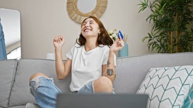 Laptop ve kredi kartıyla alışveriş yapan genç bir kadın evdeki koltukta oturmuş kutlama yapıyor.