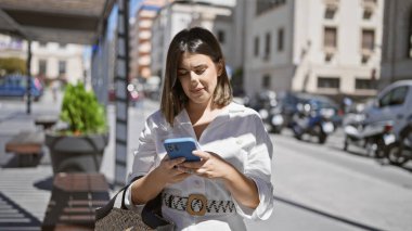 Genç, güzel, İspanyol bir kadın gülümsüyor. Kentte akıllı telefon kullanıyor.