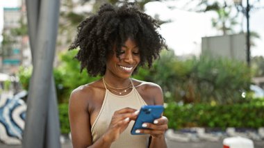 Parkta gülümseyen akıllı telefon kullanan Afrikalı Amerikalı kadın.