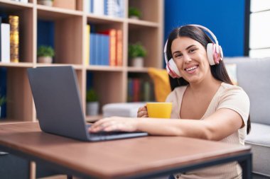 Young beautiful hispanic woman using laptop drinking coffee sitting on floor at home