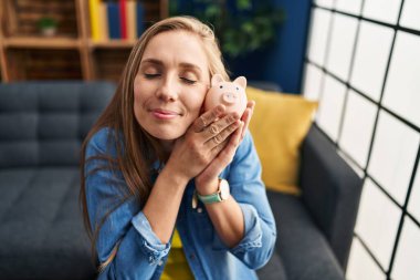 Kumbarayı elinde tutan genç sarışın kadın evdeki kanepede oturuyor.