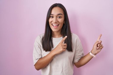 Young hispanic woman standing over pink background smiling and looking at the camera pointing with two hands and fingers to the side. 