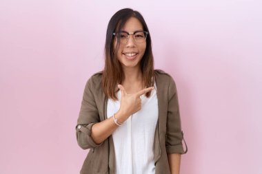 Orta yaşlı Çinli bir kadın pembe arka planda gözlük takıyor. Yüzünde mutlu ve doğal bir ifadeyle elleri ve parmaklarıyla gülümsüyor. 