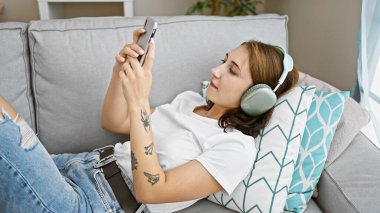 Akıllı telefon kullanan, kulaklık takan, evdeki kanepede yatan genç bir kadın.