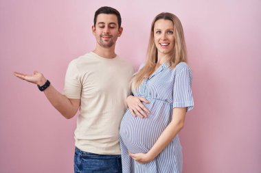 Genç bir çift pembe arka planda duran bir bebek bekliyor. Elleri açık, fotokopi alanını gösteren avuç içleri açık, gülümseyerek mutlu bir gülümseme sergiliyorlar. 