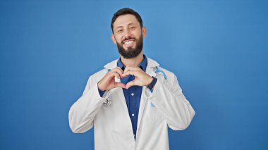 Genç İspanyol doktor gülümsüyor kalp hareketi yapıyor elleri izole edilmiş mavi arka planda.
