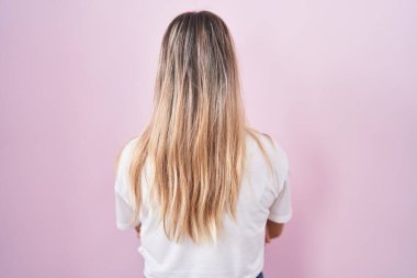 Young blonde woman standing over pink background standing backwards looking away with crossed arms 