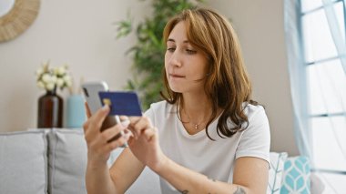 Akıllı telefon ve kredi kartıyla alışveriş yapan genç bir kadın evdeki kanepede oturuyor.