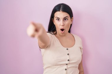 Young hispanic woman standing over pink background pointing with finger surprised ahead, open mouth amazed expression, something on the front 