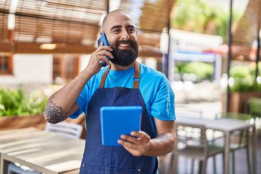Genç, kel garson, kafenin terasında dokunmatik ped kullanarak akıllı telefondan konuşuyor.