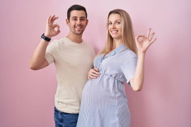 Pembe arka planda bebek bekleyen genç bir çift olumlu gülümsüyor ve el ve parmaklarıyla 