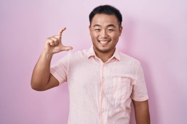 Pembe arka planda duran Çinli genç adam gülümsüyor ve el hareketleriyle hareket ediyor parmaklarıyla işaret veriyor ve kameraya bakıyor. ölçüm kavramı. 
