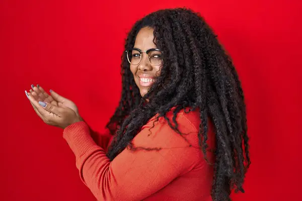 コピースペースを示す手の開いた手のひらを指す赤い背景の上に立っているプラスサイズのヒスパニック女性 興奮する笑顔の広告を提示 — ストック写真