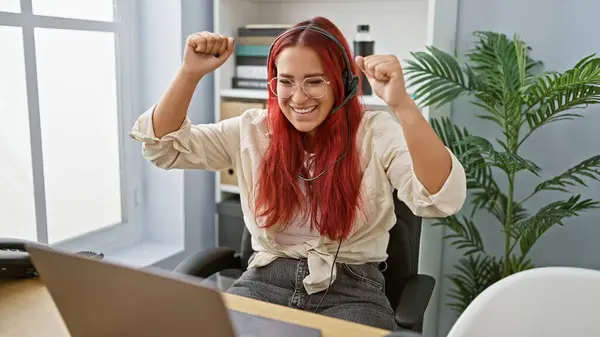 Beaming redhead business woman celebrating work victory indoors, young professional elatedly using laptop and headphones in office