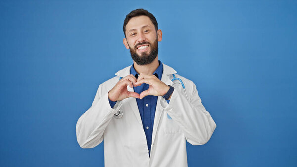 Young hispanic man doctor smiling doing heart gesture with hands over isolated blue background