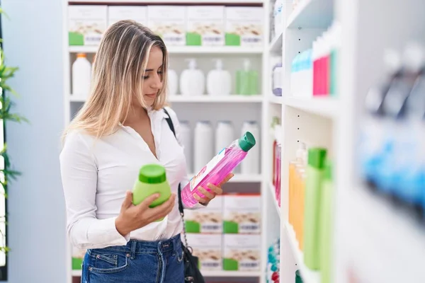 Young blonde woman customer smiling confident holding shampoo bottles at pharmacy