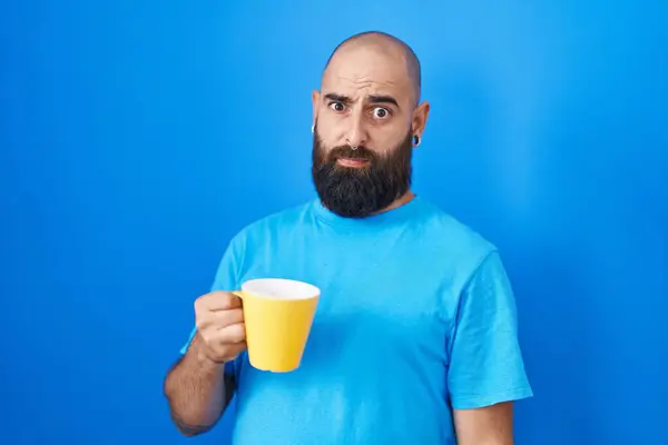 Young hispanic man with beard and tattoos drinking a cup of coffee in shock face, looking skeptical and sarcastic, surprised with open mouth