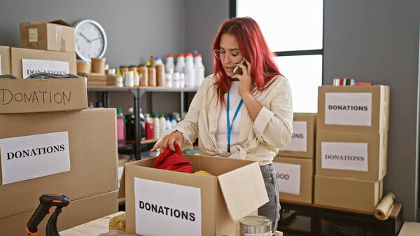 Сострадательная молодая рыжая женщина, добровольно вызвавшаяся в благотворительный центр, душевно балансируя телефонный разговор и коробку одежды, чтобы пожертвовать