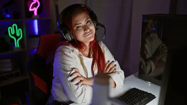 リラックスした若い赤毛の女性ストリーマー 腕が交差し 彼女のゲームルームで未来的なビデオゲームストリーミングに没頭しながら笑顔をフラッシュし デジタルエンターテインメントでサイバー夜を栽培 — ストック写真