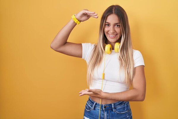 Молодая блондинка, стоящая на жёлтом фоне в наушниках, жестикулирует руками, показывая большой и большой знак, измеряющий символ. улыбаясь, глядя в камеру. концепция измерения. 