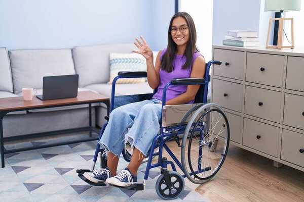 Young hispanic woman sitting on wheelchair at home showing and pointing up with fingers number four while smiling confident and happy. 